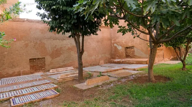 Tiled grave slabs on grass beneath fig trees, against the weathered pisé wall of the enclosure