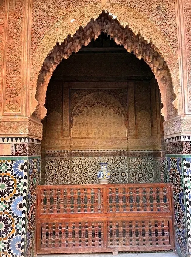 A carved cedar niche under a muqarnas arch, framed by zellige columns, with a painted ceramic vase on the wooden screen