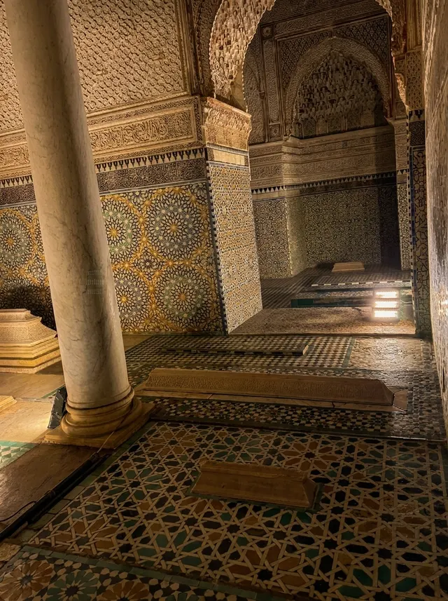 A carved marble tomb on an intricate zellige floor, lit low, with the inscribed frieze above